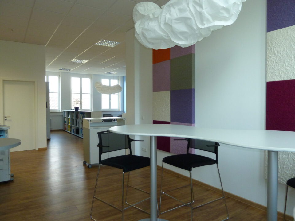 Moderner Büroraum mit Holzfußboden, weißen Wänden und großen Fenstern, einem hohen Tisch mit zwei schwarzen Stühlen im Vordergrund sowie einer wolkenförmigen Leuchte und farbigen Wandpaneelen im Hintergrund.