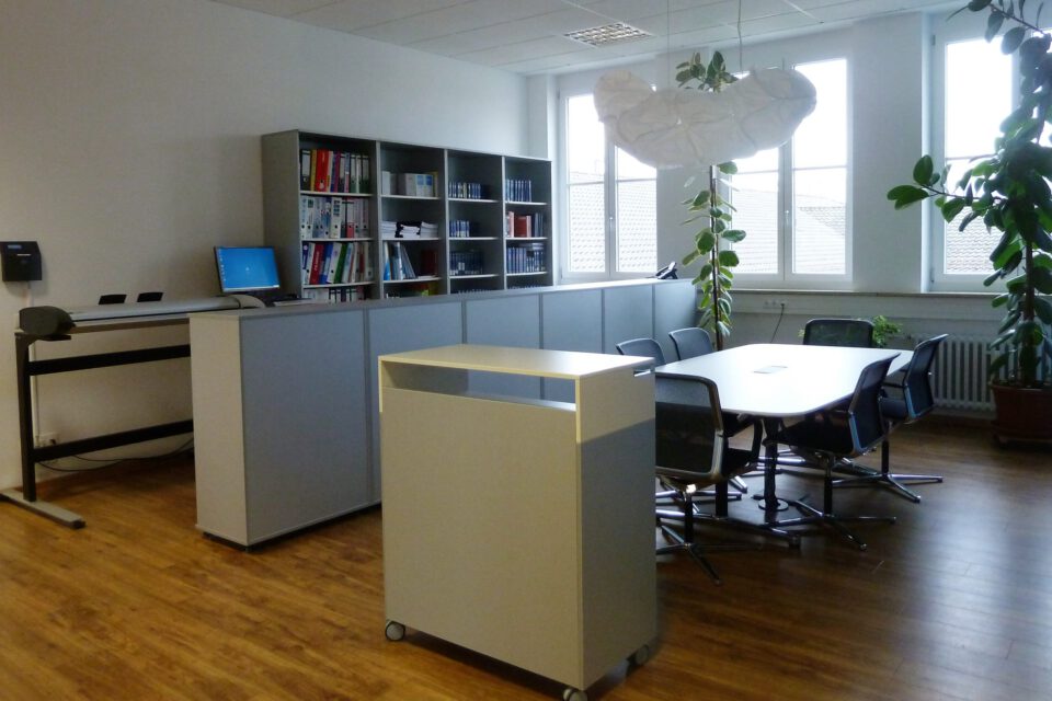 Moderner Besprechungsraum mit großem Tisch und Stühlen, Holzfußboden, Regalen mit Büchern und Ordnern im Hintergrund, einem Schreibtisch mit Computer sowie großen Fenstern und mehreren Pflanzen.