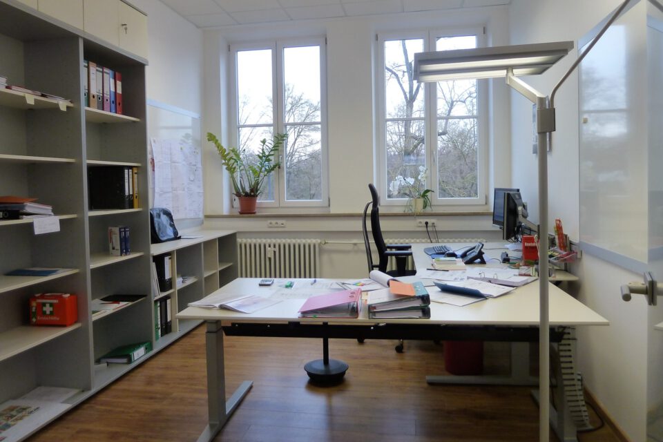 Büro mit zentralem Schreibtisch, bedeckt mit Unterlagen, Ordnern und Büromaterial, einem Monitor rechts, Regalen mit Büchern und Ordnern links sowie großen Fenstern mit Blick ins Grüne und mehreren Pflanzen.