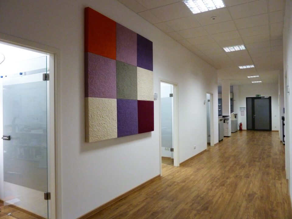 Moderner Büroflur mit Holzfußboden, weißen Wänden und farbigem Wandkunstwerk aus neun strukturierten Quadraten auf der linken Seite, mehreren Türen zu Büroräumen sowie einem Notausgang am Ende des Flurs.