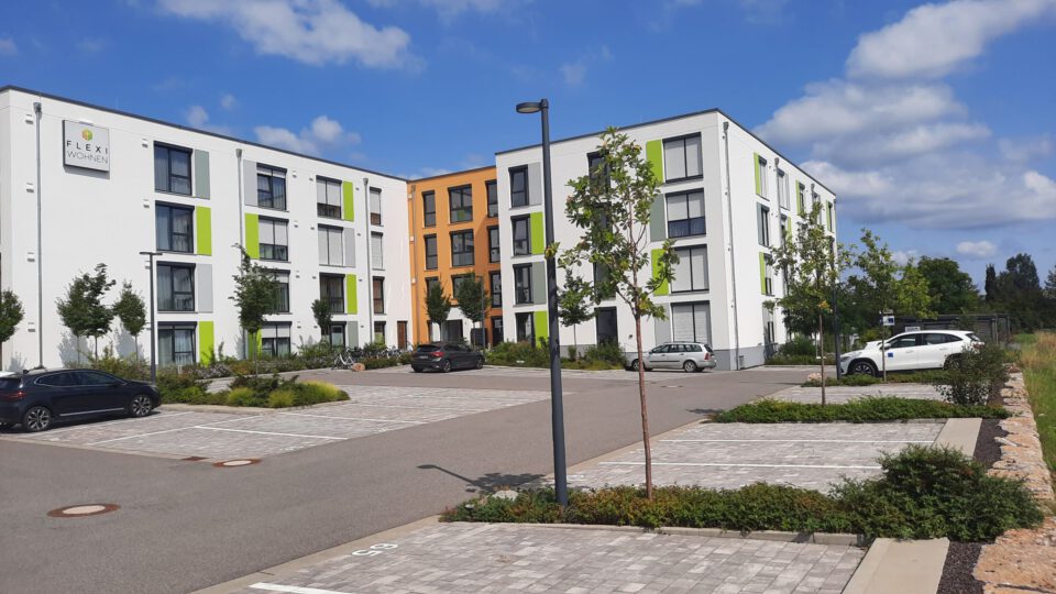 Moderner vierstöckiger Apartmentkomplex in Weiß, Grün und Orange, einem Parkplatz mit mehreren Autos im Vordergrund sowie Bepflanzung.