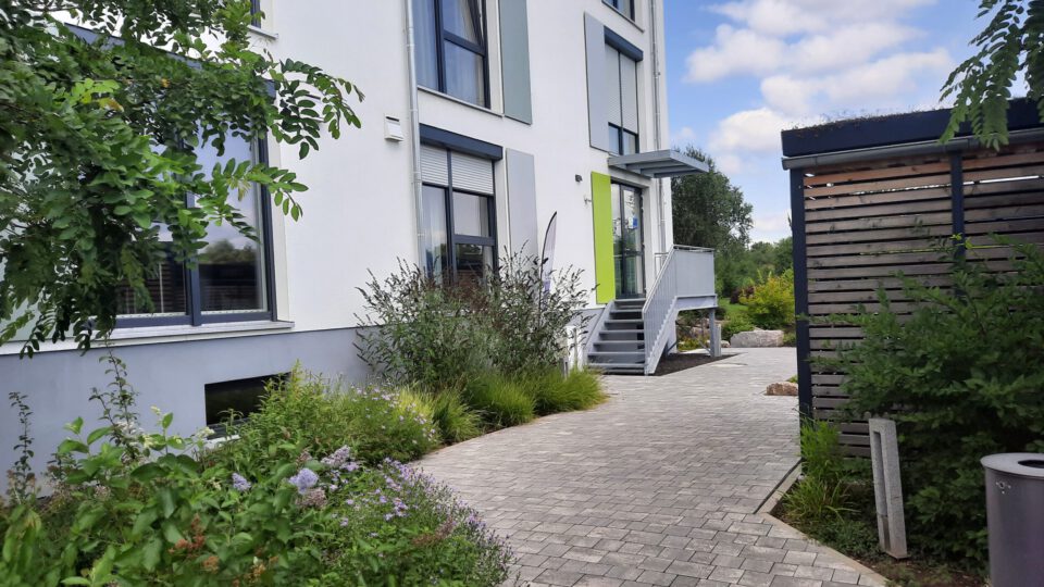 Außenansicht eines modernen Gebäudes mit weißen Wänden, großen Fenstern und grünem Eingang, erreichbar über einen gepflasterten Weg mit Treppe, umgeben von bepflanzten Grünflächen und einer Holzstruktur auf der rechten Seite.