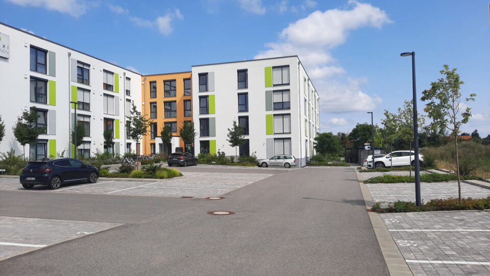 Moderner vierstöckiger Apartmentkomplex in Weiß, Grün und Orange, einem Parkplatz mit mehreren Autos im Vordergrund sowie Bepflanzung.
