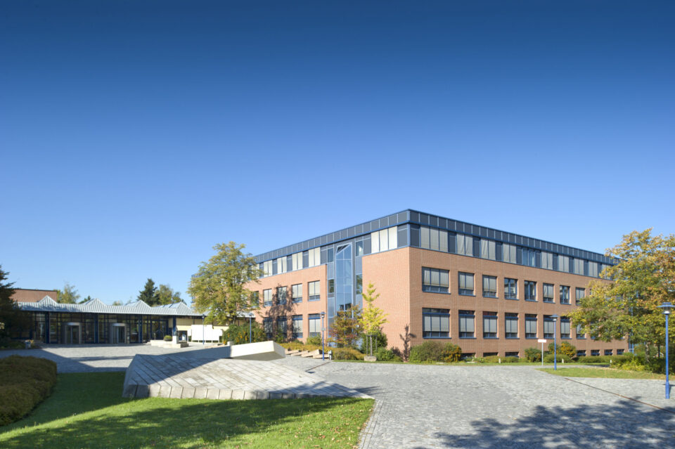 Ansicht in Richtung des Haupteingangs des LGA-Hauptgebäudes in Nürnberg, umgeben von Bäumen und Grünflächen bei sonnigem Wetter.