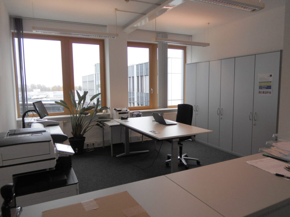 Modernes Büro mit großem Fenster, das Tageslicht hereinlässt, einem Schreibtisch mit Laptop, Bürostuhl, mehreren Schränken und Druckern sowie einer Zimmerpflanze in Fensternähe.