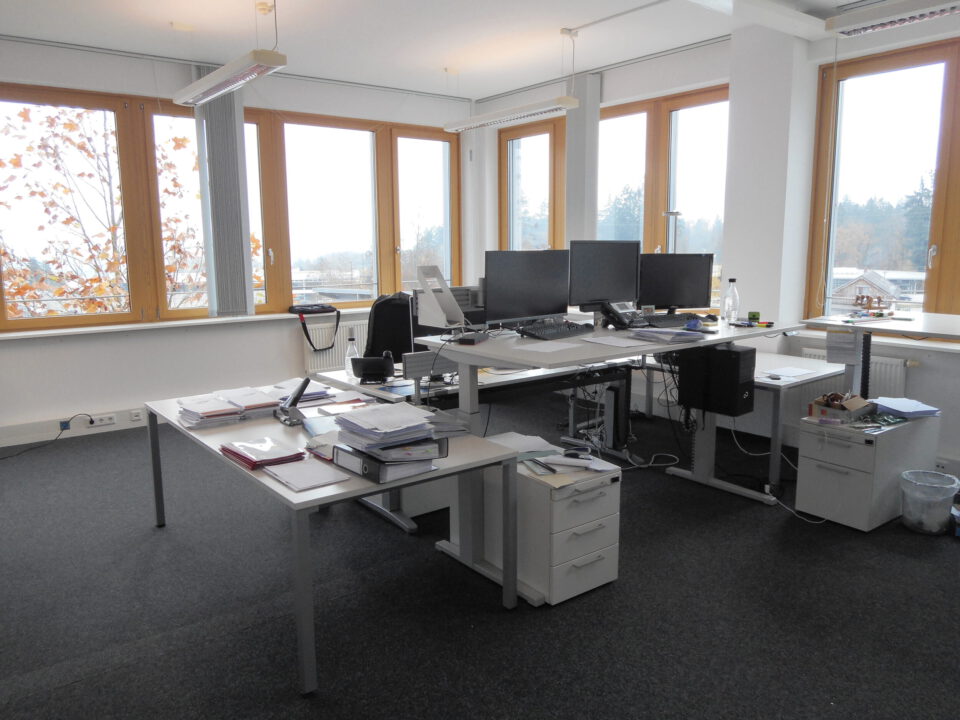 Modernes Büro mit mehreren Schreibtischen und Monitoren, großen Fenstern auf zwei Seiten für viel Tageslicht, Büroausstattung wie Tastaturen und Unterlagen sowie Teppichboden und einem Papierkorb in der Ecke.