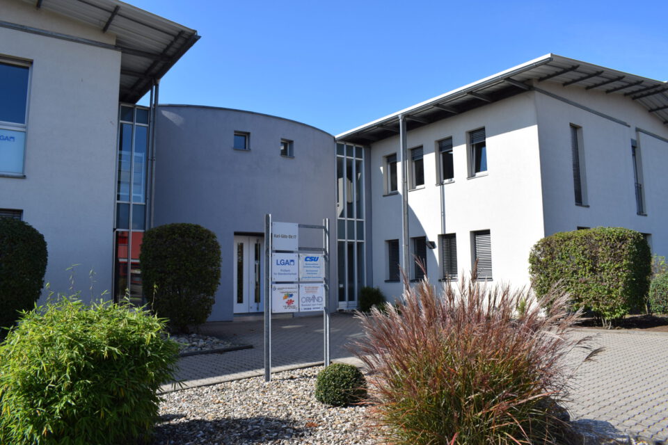 Moderner Bürokomplex mit zwei Gebäuden in zeitgemäßem Design, großen Fenstern und flachen Dächern, einem Firmenschild mit Logos wie „LGA“ und „CSU“ im Eingangsbereich sowie umliegender Begrünung bei sonnigem Wetter.