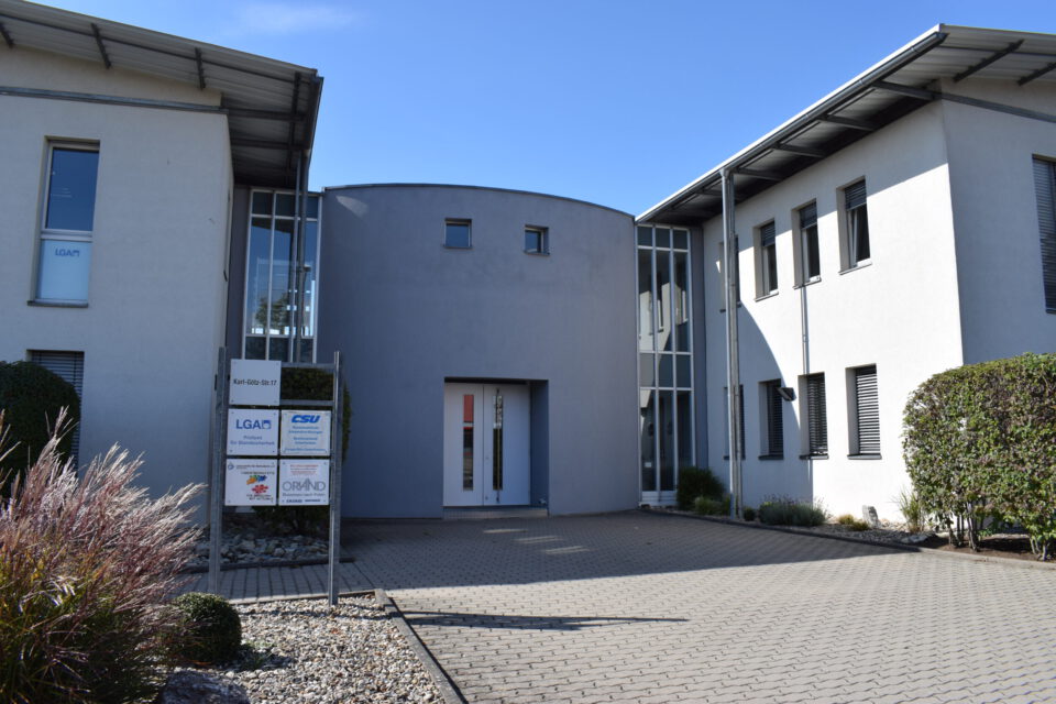 Haupteingang eines modernen Bürokomplexes mit zwei Gebäuden in zeitgemäßem Design, großen Fenstern und flachen Dächern, einem Firmenschild mit Logos wie „LGA“ und „CSU“ im Eingangsbereich sowie umliegender Begrünung bei sonnigem Wetter.