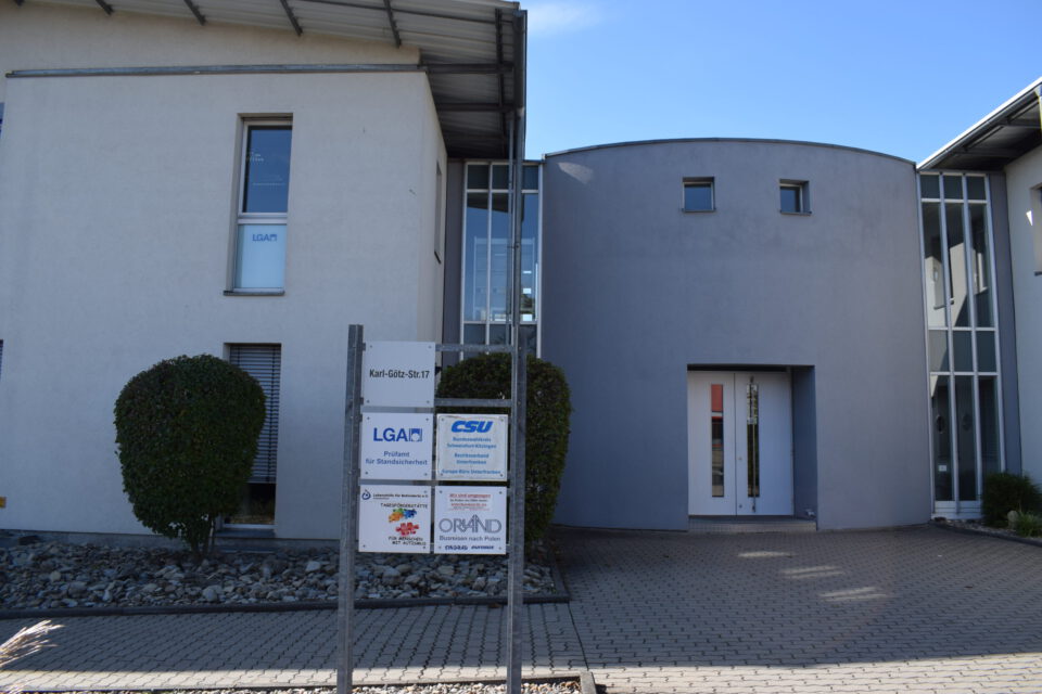 Haupteingang eines modernen Bürokomplexes mit zwei Gebäuden in zeitgemäßem Design, großen Fenstern und flachen Dächern, einem Firmenschild mit Logos wie „LGA“ und „CSU“ im Eingangsbereich bei sonnigem Wetter.