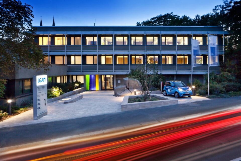 Abendaufnahme eines modernen Bürogebäudes mit großen, beleuchteten Fenstern, einem „LGA“-Schild im Vordergrund, einem kleinen Elektroauto nahe dem Eingang sowie gepflegtem Weg, Begrünung und Kirchtürmen im Hintergrund.