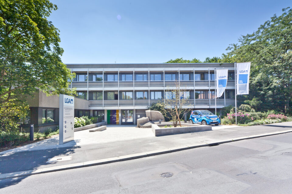 Modernes, dreistöckiges Gebäude mit Flachdach und großen Fenstern, einem gepflasterten Vorplatz mit Bepflanzung, zwei Fahnen mit „LGA“-Logo, einem LGA-Schild am Eingang sowie einem blauen Auto im Vordergrund.
