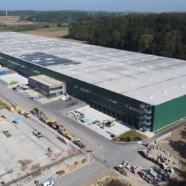 Das Bild zeigt ein großes Industrie- oder Logistikzentrum mit grüner Fassade, flachem Dach, mehreren Laderampen, parkenden Lkw sowie umliegenden Baufahrzeugen, Materialien und einer Mischung aus befestigten Flächen und unbebautem Gelände mit Bäumen im Hintergrund.