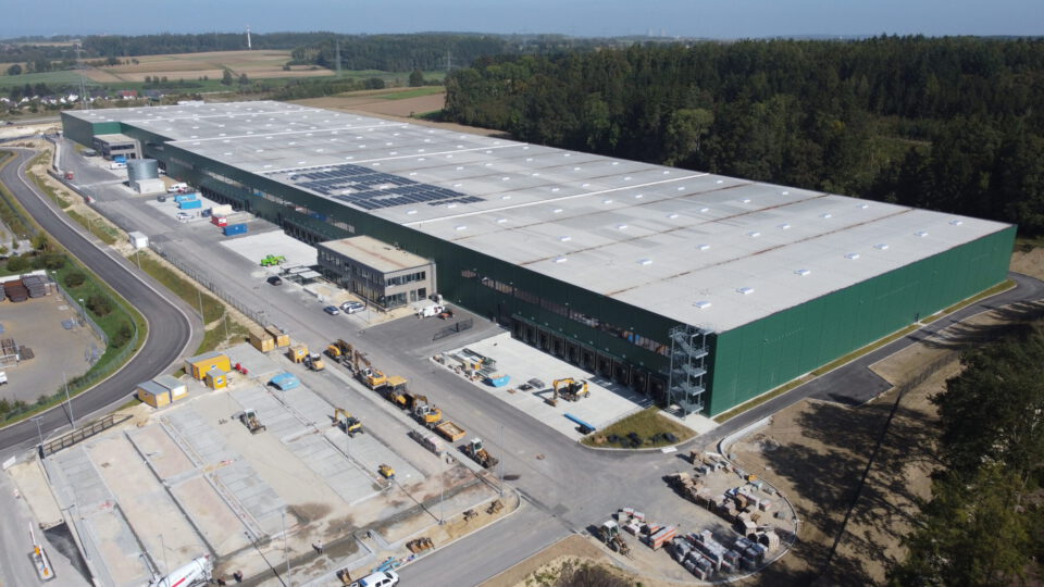 Das Bild zeigt ein großes Industrie- oder Logistikzentrum mit grüner Fassade, flachem Dach, mehreren Laderampen, parkenden Lkw sowie umliegenden Baufahrzeugen, Materialien und einer Mischung aus befestigten Flächen und unbebautem Gelände mit Bäumen im Hintergrund.