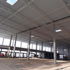 Das Bild zeigt das Innere eines großen, unfertigen Industrie- oder Gewerbebaus mit hoher Decke, Metallträgern, Oberlichtern, Betonstützen, einer Galerie mit Geländer sowie Baustellenfahrzeugen und unbearbeitetem Boden.