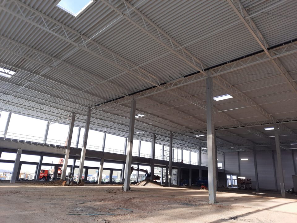 Das Bild zeigt das Innere eines großen, unfertigen Industrie- oder Gewerbebaus mit hoher Decke, Metallträgern, Oberlichtern, Betonstützen, einer Galerie mit Geländer sowie Baustellenfahrzeugen und unbearbeitetem Boden.