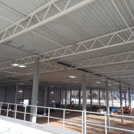 Das Bild zeigt das Innere eines großen, teilweise errichteten Industrie- oder Lagergebäudes mit hoher Decke, Metallträgern, Betonstützen, verstreutem Baumaterial und Maschinen sowie natürlichem Lichteinfall durch Fenster auf der gegenüberliegenden Seite.