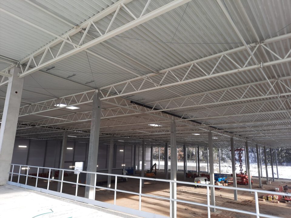 Das Bild zeigt das Innere eines großen, teilweise errichteten Industrie- oder Lagergebäudes mit hoher Decke, Metallträgern, Betonstützen, verstreutem Baumaterial und Maschinen sowie natürlichem Lichteinfall durch Fenster auf der gegenüberliegenden Seite.