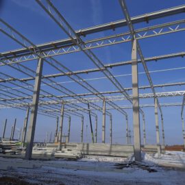 Das Bild zeigt den Rohbau eines Stahltragwerks mit vertikalen und horizontalen Trägern sowie Dachträgern, umgeben von gestapeltem Baumaterial auf schneebedecktem Boden unter klarem, blauem Himmel.