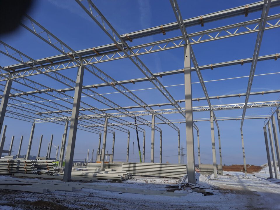 Das Bild zeigt den Rohbau eines Stahltragwerks mit vertikalen und horizontalen Trägern sowie Dachträgern, umgeben von gestapeltem Baumaterial auf schneebedecktem Boden unter klarem, blauem Himmel.