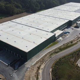 Das Bild zeigt einen großen Industriekomplex mit mehreren verbundenen Hallen mit weißen Dächern und grünen Wänden, mehreren Laderampen, einer Zufahrtsstraße mit Baufahrzeugen sowie umliegendem Grün und einem benachbarten Gebäude mit Solarpanels auf dem Dach.