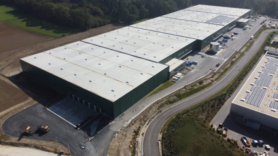 Das Bild zeigt einen großen Industriekomplex mit mehreren verbundenen Hallen mit weißen Dächern und grünen Wänden, mehreren Laderampen, einer Zufahrtsstraße mit Baufahrzeugen sowie umliegendem Grün und einem benachbarten Gebäude mit Solarpanels auf dem Dach.