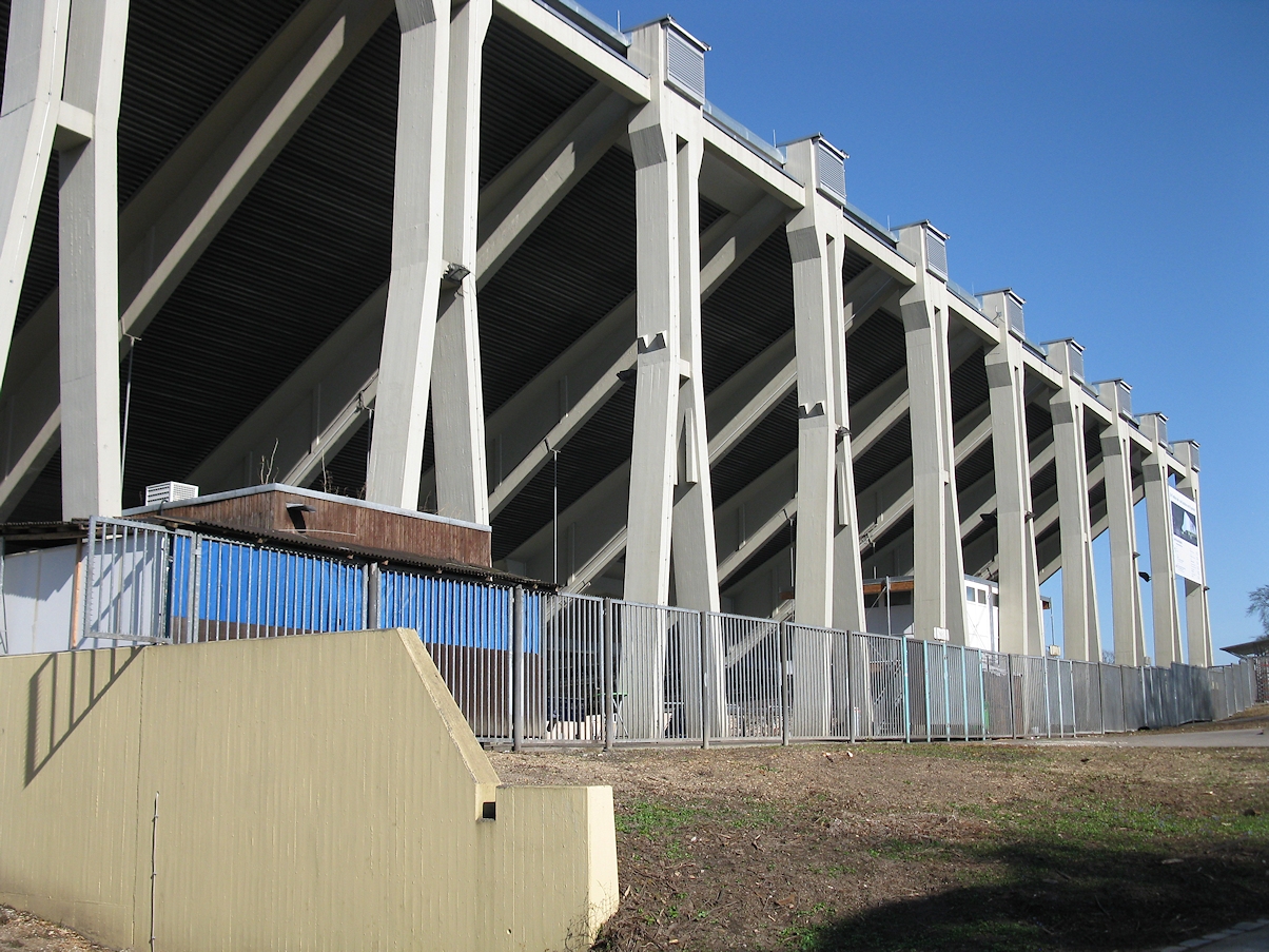 Das Bild zeigt die Außenansicht eines großen Stadions mit mehreren Betonstützen und Trägern. Hohe, senkrechte Pfeiler tragen den oberen Tribünenbereich, im unteren Bereich ist ein Metallzaun mit blauen Paneelen dahinter zu sehen.