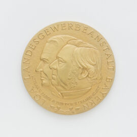 Goldene Medaille auf weißem Hintergrund mit dem geprägten Profil zweier Köpfe in der Mitte, umgeben von der Inschrift „LGA LANDESGEWERBEANSTALT BAYERN“ und „LOTHAR V. FABER & THEODOR V. CRAMER-KLETT“.