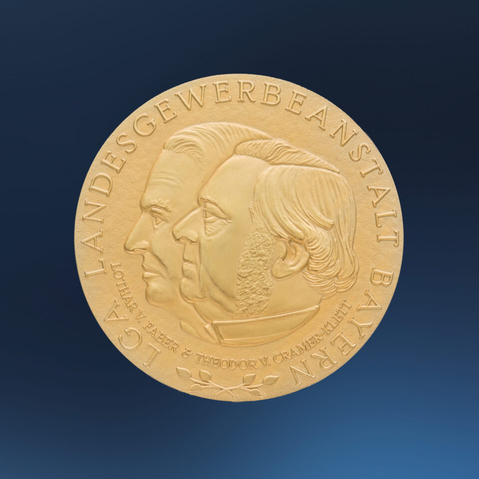 Goldene Medaille auf blauem Hintergrund mit dem geprägten Profil zweier Köpfe in der Mitte, umgeben von der Inschrift „LGA LANDESGEWERBEANSTALT BAYERN“ und „LOTHAR V. FABER & THEODOR V. CRAMER-KLETT“.