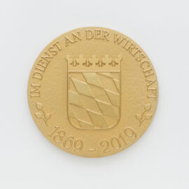 Goldene Münze mit geprägtem bayrischem Wappen auf weißem Hintergrund, das diagonale Streifen und eine Krone zeigt, umgeben von der Inschrift „IM DIENST AN DER WIRTSCHAFT“ und den Jahreszahlen „1869 – 2019“.