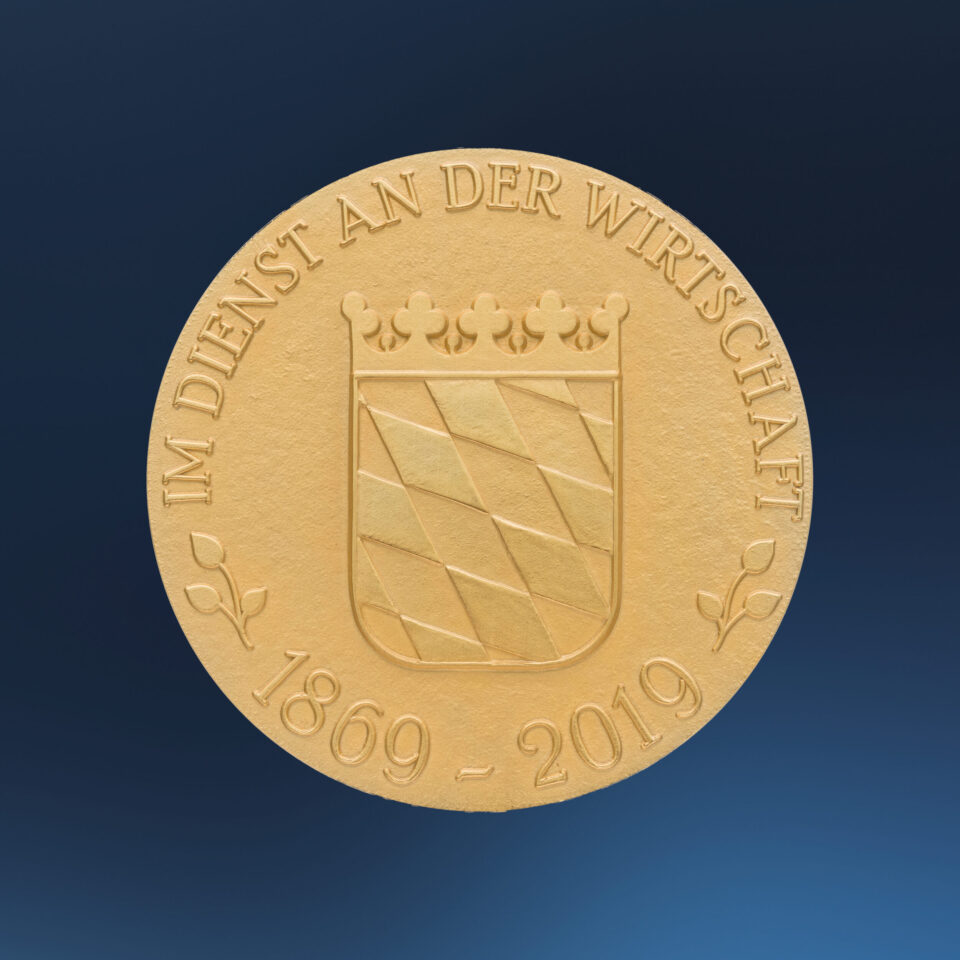 Goldene Medaille mit geprägtem bayrischem Wappen auf blauem Hintergrund, das diagonale Streifen und eine Krone zeigt, umgeben von der Inschrift „IM DIENST AN DER WIRTSCHAFT“ und den Jahreszahlen „1869 – 2019“.