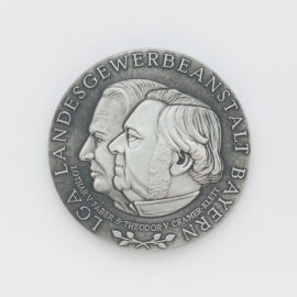 Silberne Medaille auf weißem Hintergrund mit dem geprägten Profil zweier Köpfe in der Mitte, umgeben von der Inschrift „LGA LANDESGEWERBEANSTALT BAYERN“ und „LOTHAR V. FABER & THEODOR V. CRAMER-KLETT“.