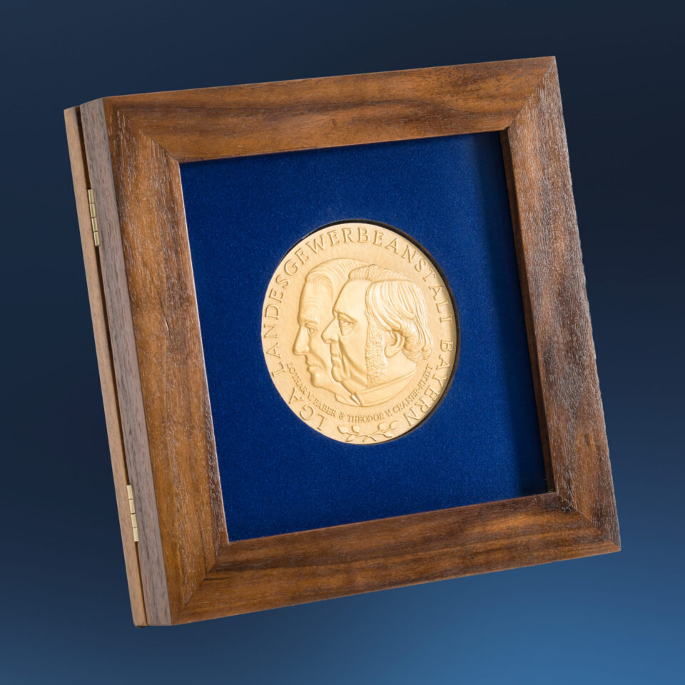 Gerahmte goldene Medaille auf blauem Hintergrund mit zwei geprägten Profilen vor blauem Hintergrund, eingefasst in einem Holzrahmen, mit der Inschrift „LGA LANDESGEWERBEANSTALT BAYERN“.