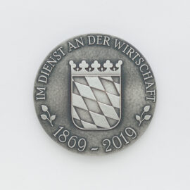 Silberne Medaille mit geprägtem bayrischem Wappen auf weißem Hintergrund, das diagonale Streifen und eine Krone zeigt, umgeben von der Inschrift „IM DIENST AN DER WIRTSCHAFT“ und den Jahreszahlen „1869 – 2019“.
