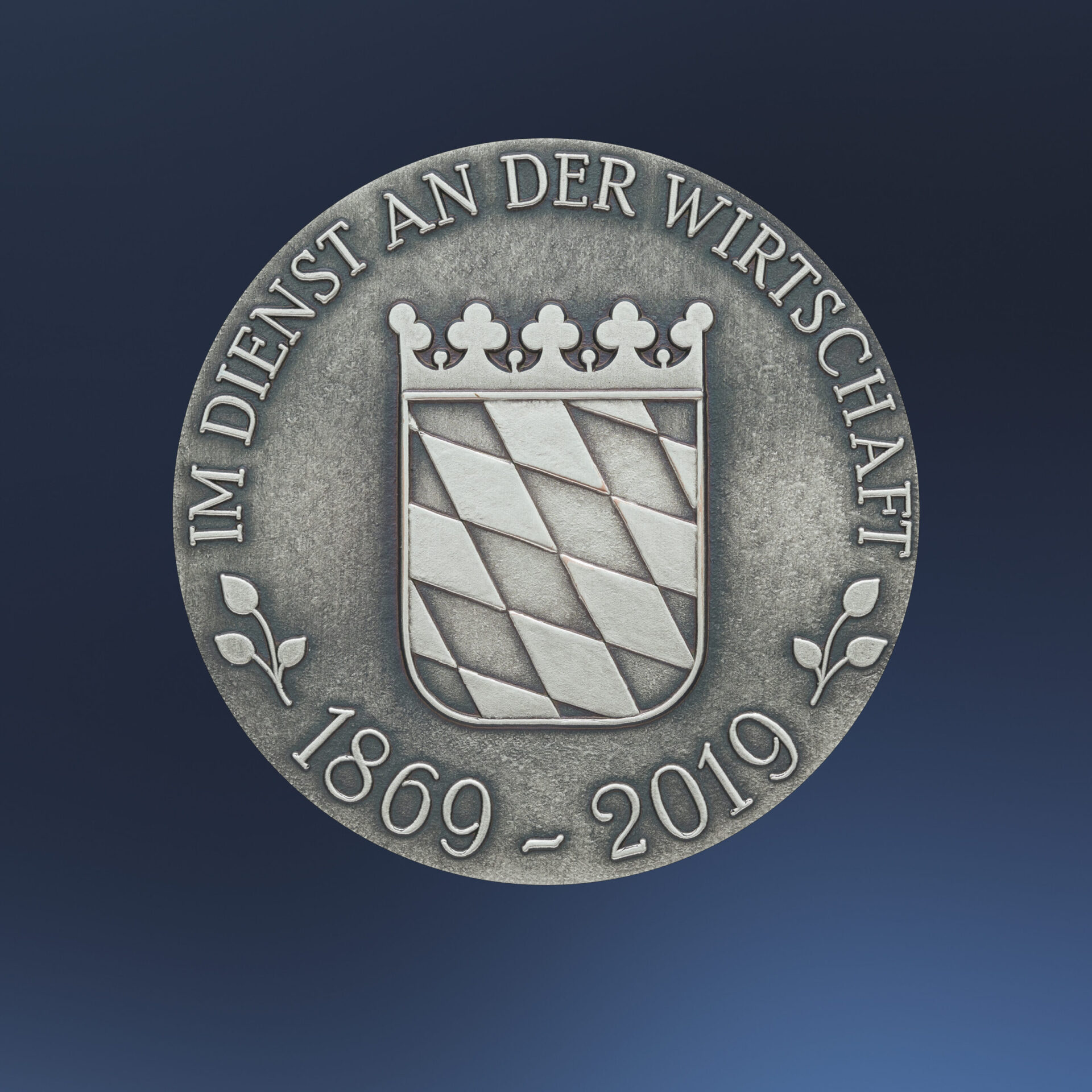 Silberne Medaille mit geprägtem bayrischem Wappen auf blauem Hintergrund, das diagonale Streifen und eine Krone zeigt, umgeben von der Inschrift „IM DIENST AN DER WIRTSCHAFT“ und den Jahreszahlen „1869 – 2019“.