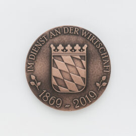 Bronzene Medaille mit geprägtem bayrischem Wappen auf weißem Hintergrund, das diagonale Streifen und eine Krone zeigt, umgeben von der Inschrift „IM DIENST AN DER WIRTSCHAFT“ und den Jahreszahlen „1869 – 2019“.
