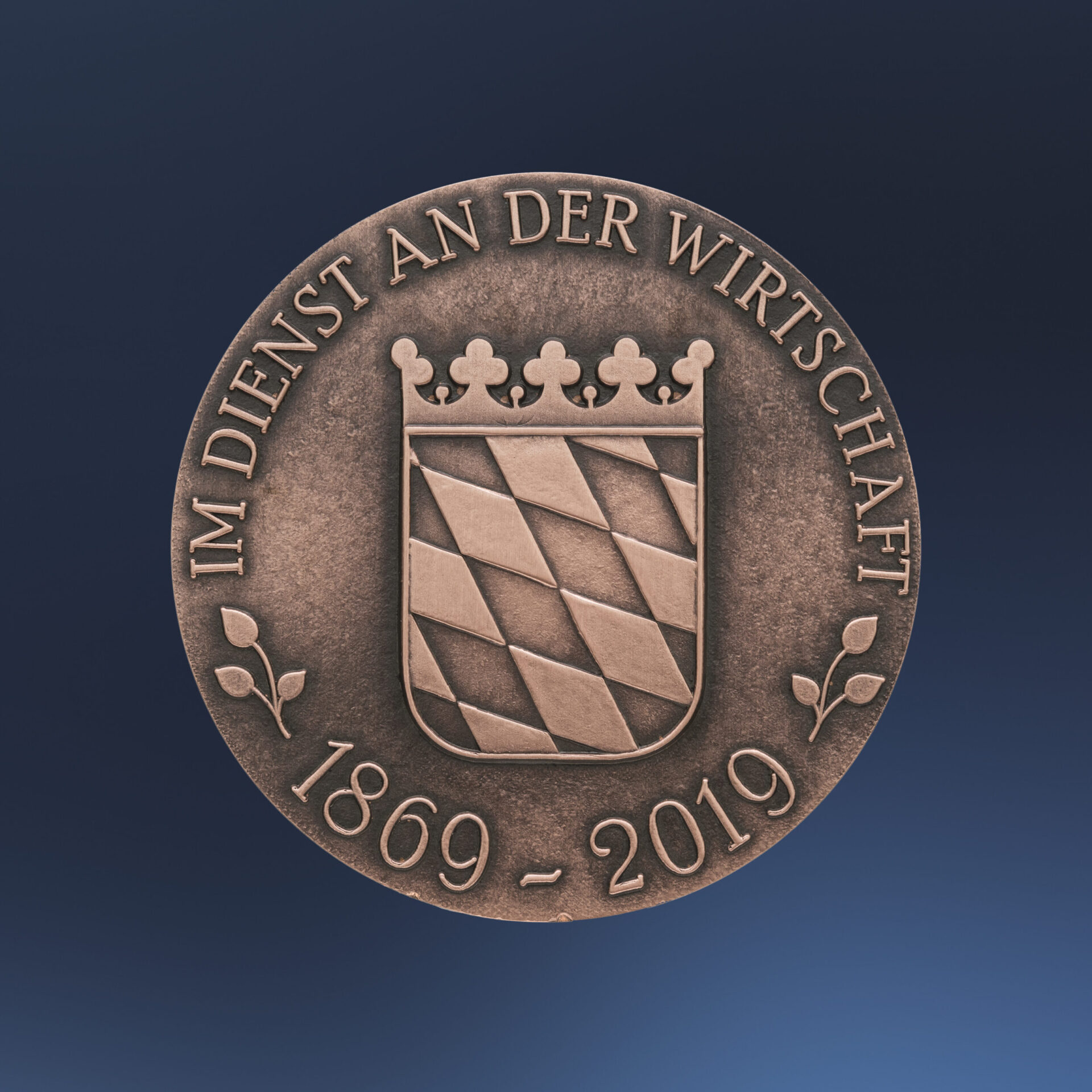 Bronzene Medaille mit geprägtem bayrischem Wappen auf blaum Hintergrund, das diagonale Streifen und eine Krone zeigt, umgeben von der Inschrift „IM DIENST AN DER WIRTSCHAFT“ und den Jahreszahlen „1869 – 2019“.
