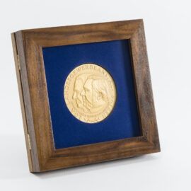 Gerahmte goldene Medaille mit zwei geprägten Profilen vor blauem Hintergrund, eingefasst in einem Holzrahmen, mit der Inschrift „LGA LANDESGEWERBEANSTALT BAYERN“.