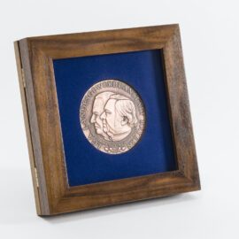 Gerahmte bronzene Medaille mit zwei geprägten Profilen vor blauem Hintergrund, eingefasst in einem Holzrahmen, mit der Inschrift „LGA LANDESGEWERBEANSTALT BAYERN“.