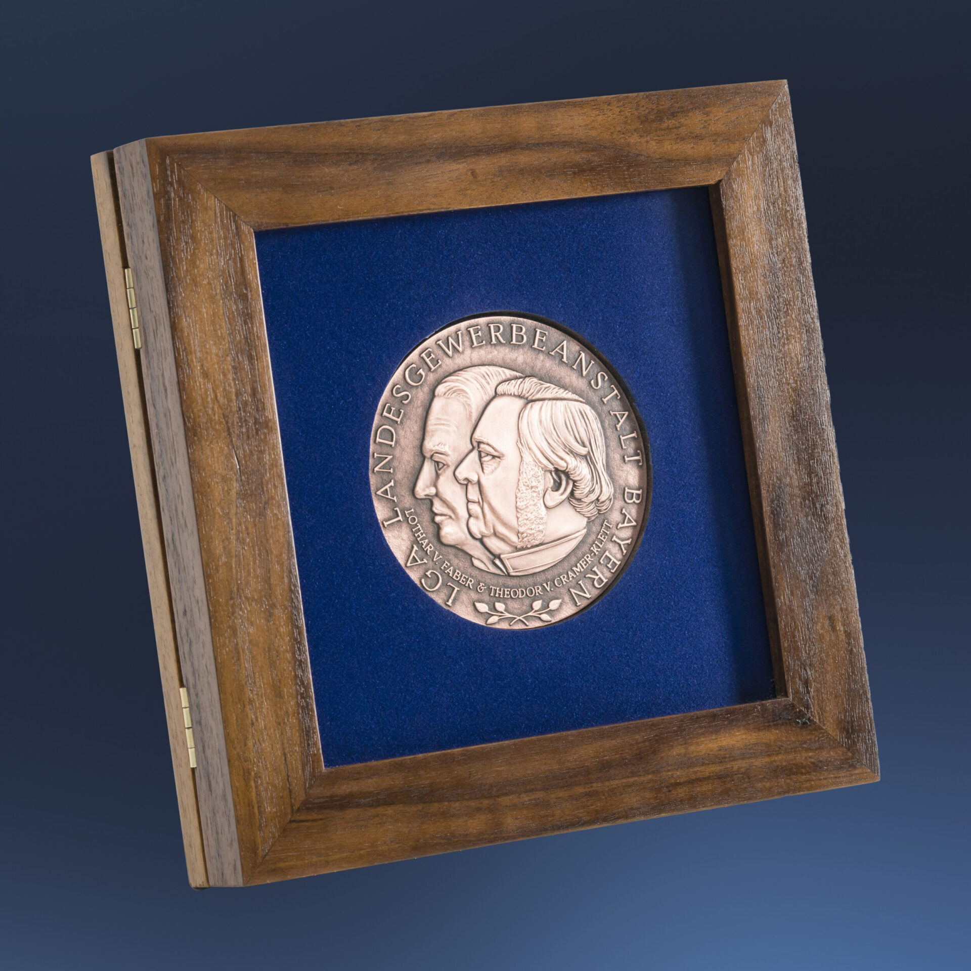 Gerahmte bronzene Medaille mit zwei geprägten Profilen vor blauem Hintergrund, eingefasst in einem Holzrahmen, mit der Inschrift „LGA LANDESGEWERBEANSTALT BAYERN“.