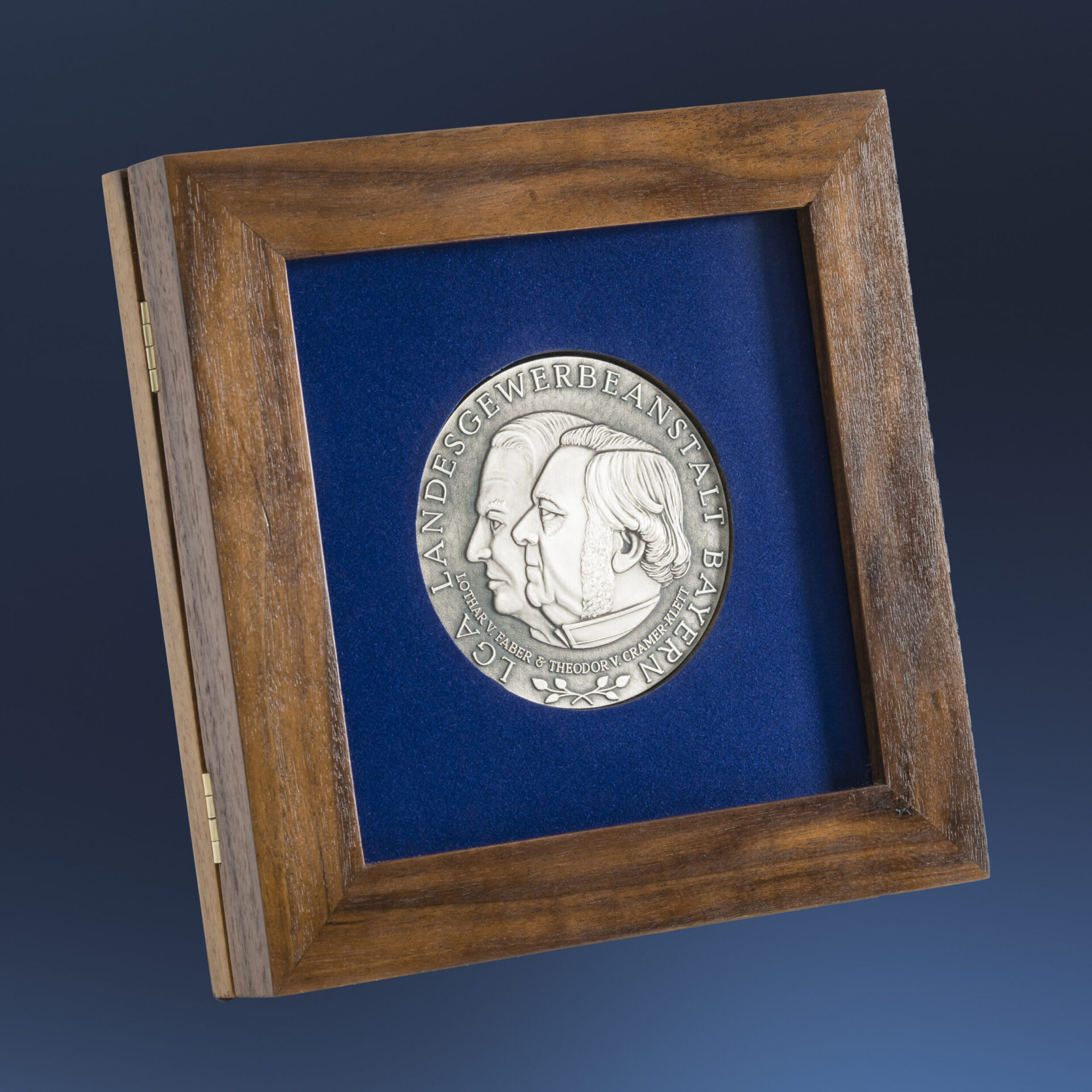 Gerahmte silberne Medaille mit zwei geprägten Profilen vor blauem Hintergrund, eingefasst in einem Holzrahmen, mit der Inschrift „LGA LANDESGEWERBEANSTALT BAYERN“.