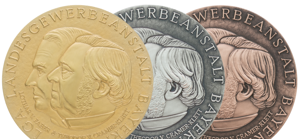 Drei nebeneinander liegende Medaillen in Gold, Silber und Bronze mit zwei geprägten Profilen und der Inschrift „LANDESGEWERBEANSTALT BAYERN“ am Rand.