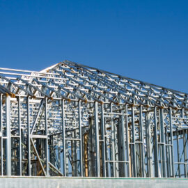 Das Bild zeigt das Metallgerüst eines Gebäudes im Bau, bestehend aus zahlreichen Stahlträgern und -stützen. Der klare blaue Himmel im Hintergrund deutet auf gutes Wetter hin.