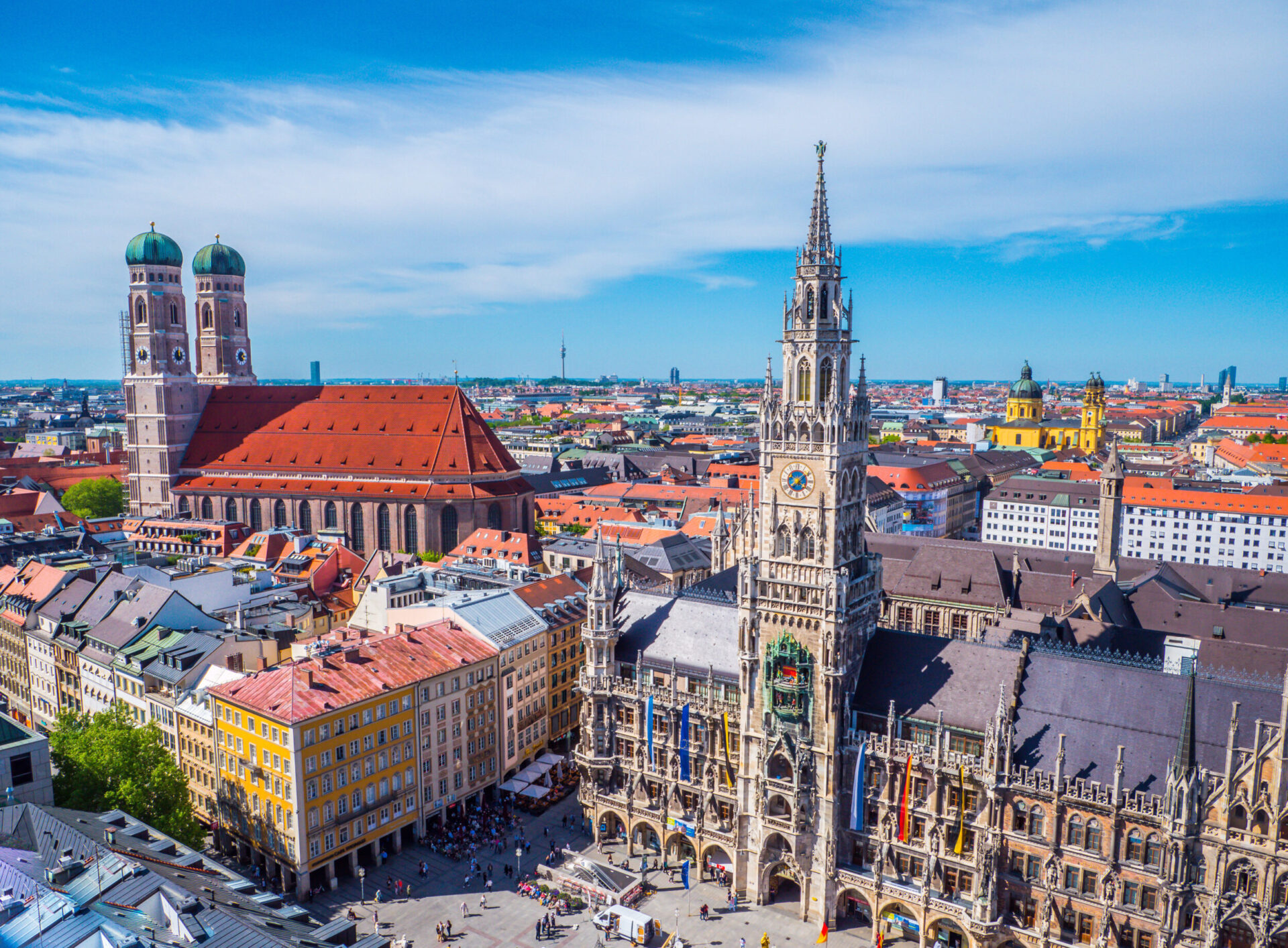 Luftaufnahme von München mit markanten Wahrzeichen wie dem Neuen Rathaus mit hohem Uhrturm und der Frauenkirche mit ihren charakteristischen Zwillingstürmen; die Stadtansicht zeigt eine Mischung aus historischen und modernen Gebäuden unter blauem Himmel.