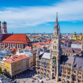 Luftaufnahme von München mit markanten Wahrzeichen wie dem Neuen Rathaus mit hohem Uhrturm und der Frauenkirche mit ihren charakteristischen Zwillingstürmen; die Stadtansicht zeigt eine Mischung aus historischen und modernen Gebäuden unter blauem Himmel.
