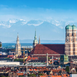 Panoramablick auf München mit der Frauenkirche und dem Neuen Rathaus im Vordergrund, dahinter die Dächer der Stadt und im Hintergrund die Alpen unter klarem Himmel.