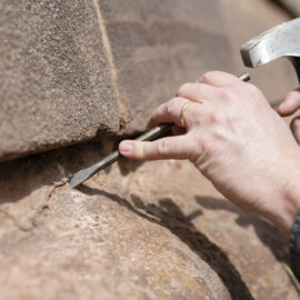 Eine Person entnimmt aus einer Sandsteinmauer eine Probe. Der Prüfer nutzt hierfür einen kleinen Hammer und Meisel.
