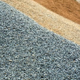 Das Bild zeigt mehrere Haufen verschiedener Baustoffe. Im Vordergrund liegt ein Haufen aus kleinem grauem Kies, dahinter befindet sich ein beiger Sandhaufen. Weiter hinten ist ein weiterer Haufen mit etwas größeren Steinen oder gebrochenem Gestein (Splitt) zu sehen.