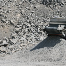 Das Bild zeigt eine Gesteinsbrechmaschine im Einsatz in einem Steinbruch. Große Steine werden zu kleineren, kiesartigen Stücken zerkleinert und über ein Förderband in einem Haufen abgelegt. Im Hintergrund ist felsiges Gelände mit unterschiedlich großen Steinen zu sehen.