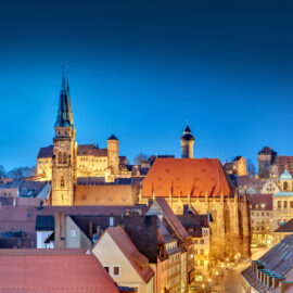 Panoramablick auf Nürnberg am Abend mit beleuchteten historischen Gebäuden, darunter Kirchen mit hohen Türmen und eine Burg mit mehreren Türmen, vor einem tiefblauen Himmel.