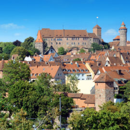 Panoramablick auf Nürnberg mit grünen Bäumen im Vordergrund, traditionellen Gebäuden mit roten Ziegeldächern in der Mitte und der historischen Kaiserburg mit mehreren Türmen und Befestigungen im Hintergrund unter blauem Himmel.